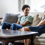 Teenaged boy sits on couch, smiling and talking to his mom